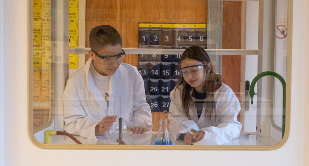 Leerlingen in de les fysica die een experiment uitvoeren met bepaalde vloeistoffen in een schort, veiligheidsbril en achter plexiglas